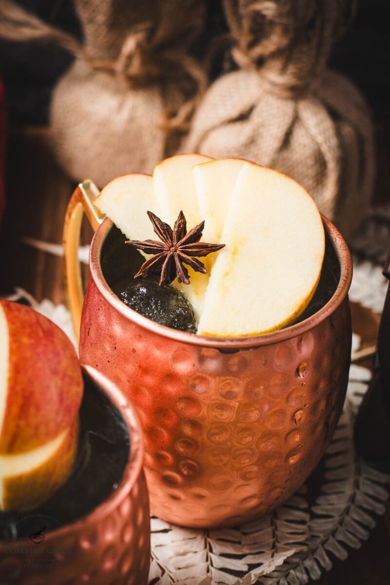 Copper mug, filled with delicious apple cider Moscow mule, garnished with fresh apple slices and a star anise. Placed on wooden board.