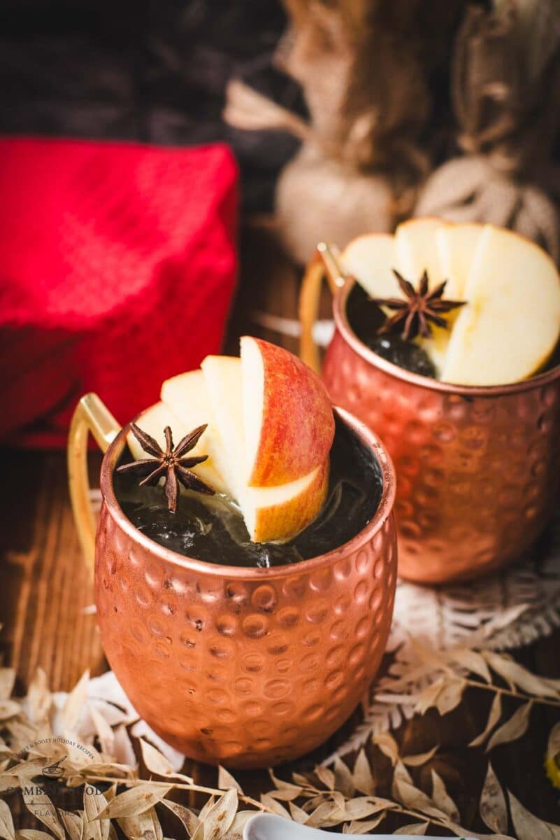 Copper mug, filled with delicious apple cider Moscow mule, garnished with fresh apple slices and a star anise. Placed on wooden board, next to a red cloth.