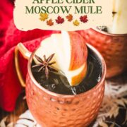 Copper mug, filled with delicious apple cider Moscow mule, garnished with fresh apple slices and a star anise. Placed on wooden board, next to a red cloth.