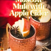 Copper mug, filled with delicious apple cider Moscow mule, garnished with fresh apple slices and a star anise. Placed on wooden board, next to a red cloth.