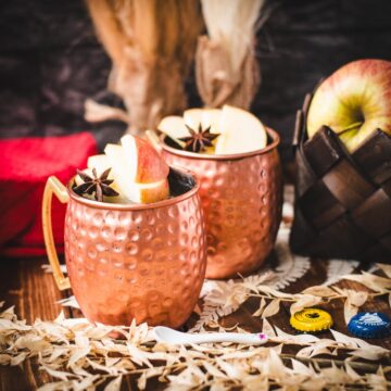 Copper mug, filled with delicious apple cider Moscow mule, garnished with fresh apple slices and a star anise. Placed on wooden board, next to a red cloth.