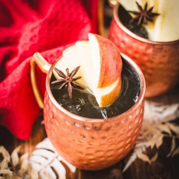 Copper mug, filled with delicious apple cider Moscow mule, garnished with fresh apple slices and a star anise. Placed on wooden board, next to a red cloth.