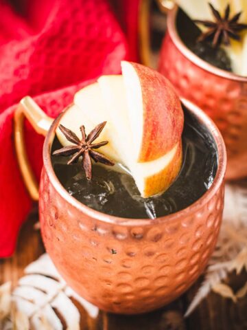 Copper mug, filled with delicious apple cider Moscow mule, garnished with fresh apple slices and a star anise. Placed on wooden board, next to a red cloth.