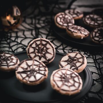 Hauntingly delicious spiderweb cookies, placed on black plate on spiderweb ground.