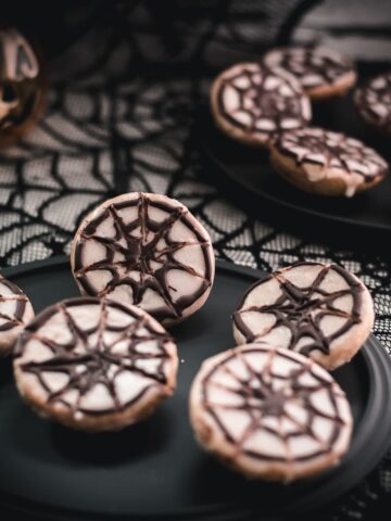 Hauntingly delicious spiderweb cookies, placed on black plate on spiderweb ground.