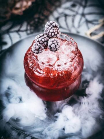 Spooky kramble cocktail, garnished with frozen blackberries and placed on a foggy plate.