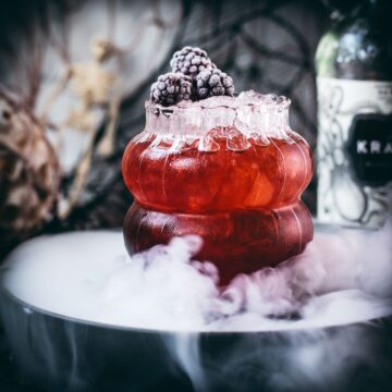 Spooky kramble cocktail, garnished with frozen blackberries and placed on a foggy plate.