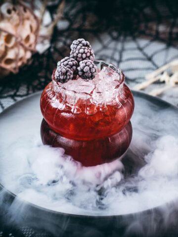 Spooky kramble cocktail, garnished with frozen blackberries and placed on a foggy plate.