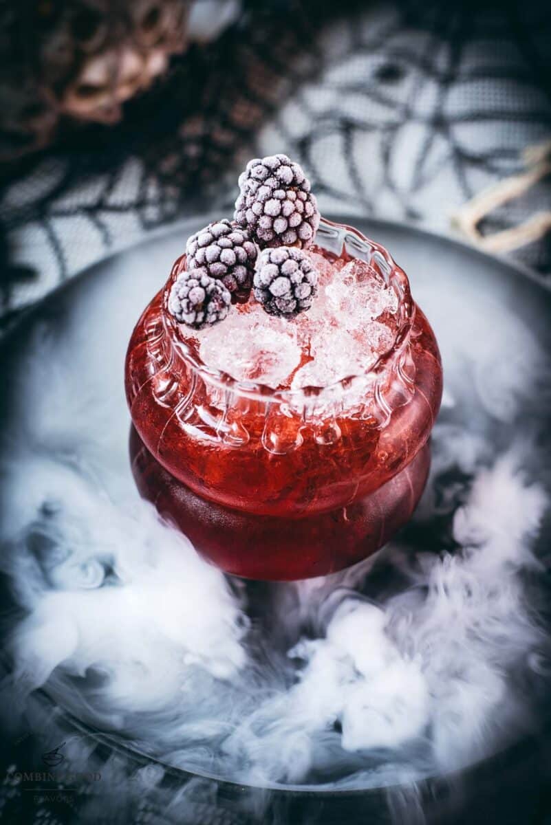 Spooky kramble cocktail, garnished with frozen blackberries and placed on a foggy plate.