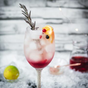 Gorgeous wineglass filled with delicious layered Prosecco cosmo. Garnished with a rosemary twig, an orange slice, and red and black currants.