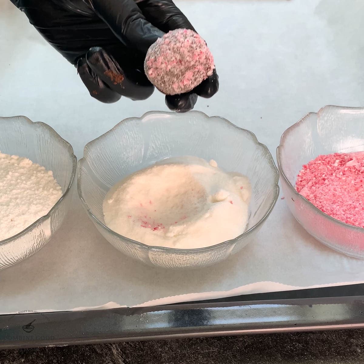 Hand with black gloves holding dough ball over a bowl filled with granulated sugar.