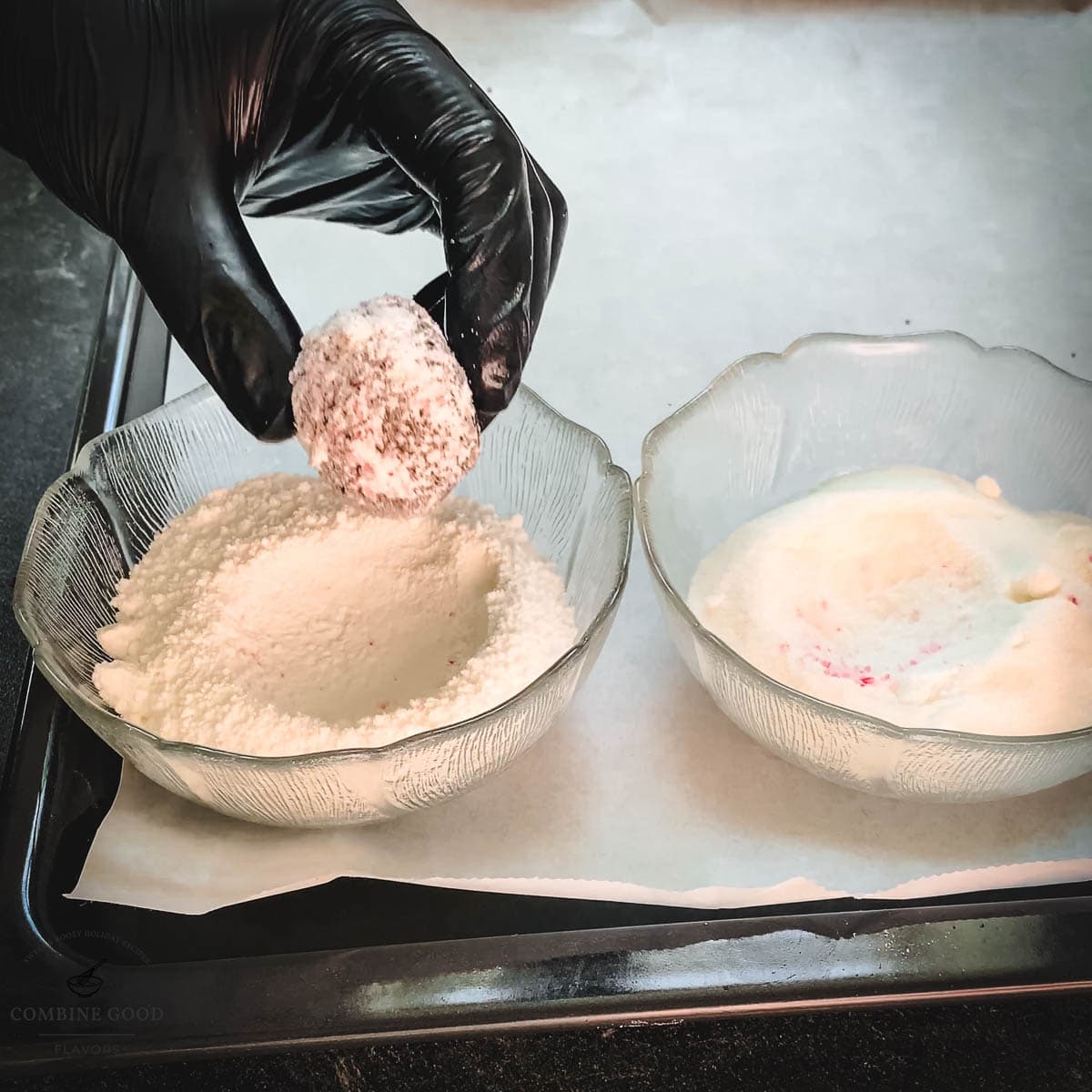 Hand with black gloves holding dough ball over a bowl filled with powdered sugar.