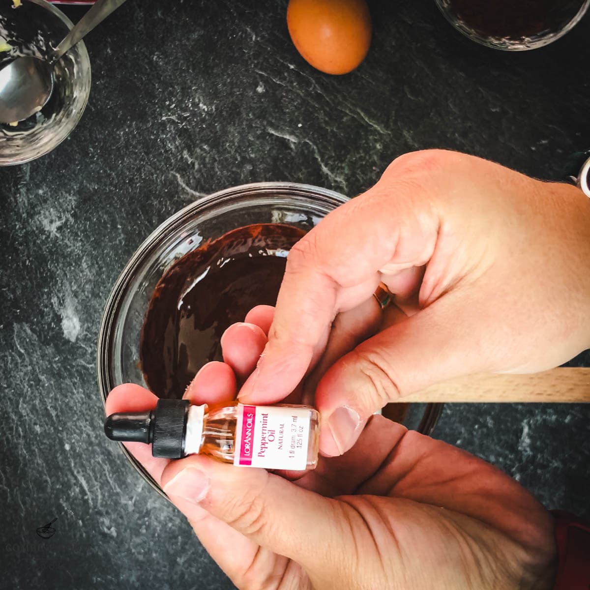 Hands holding peppermint oil over a bowl of melted chocolate.