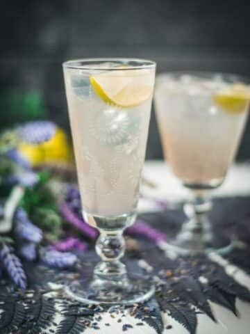 Two beautiful glasses with flower engraving filled with lavender Moscow Mule, arranged on dark blue purple leaves.