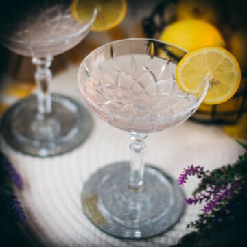 Elegant lavender gin spritz cocktail with prosecco, placed on a mirrored coaster, garnished with a lemon slice.