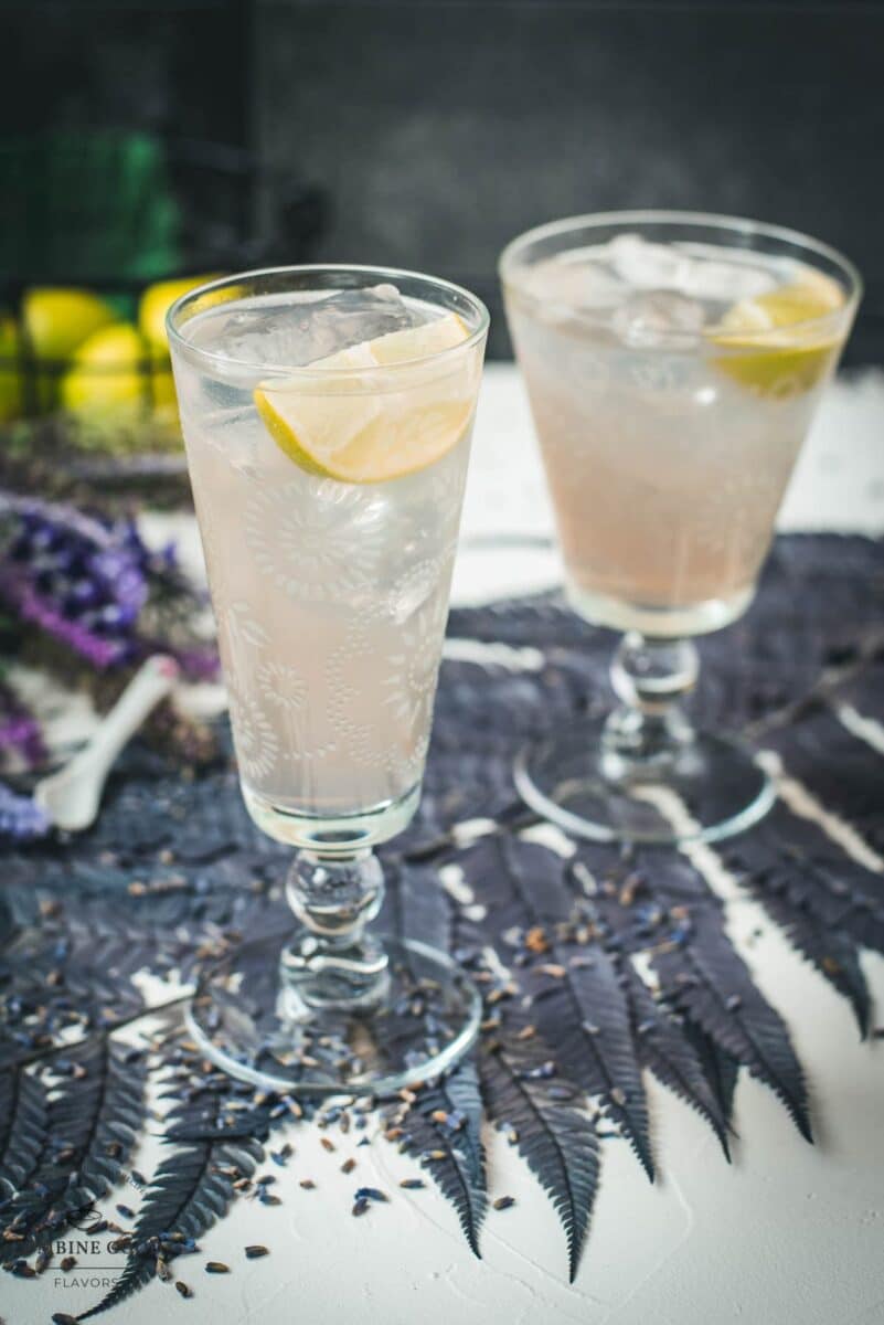 Two beautiful glasses with flower engraving filled with lavender Moscow Mule, arranged on dark blue purple leaves.