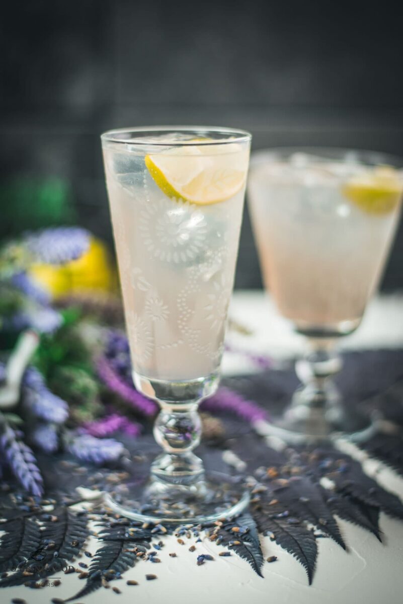 Two beautiful glasses with flower engraving filled with lavender Moscow Mule, arranged on dark blue purple leaves.