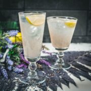 Two beautiful glasses with flower engraving filled with lavender Moscow Mule, arranged on dark blue purple leaves.