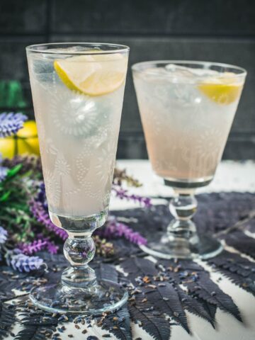 Two beautiful glasses with flower engraving filled with lavender Moscow Mule, arranged on dark blue purple leaves.