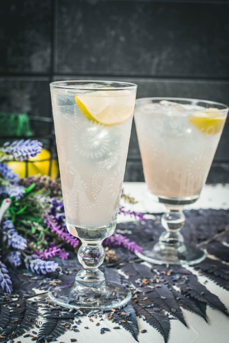 Two beautiful glasses with flower engraving filled with lavender Moscow Mule, arranged on dark blue purple leaves.