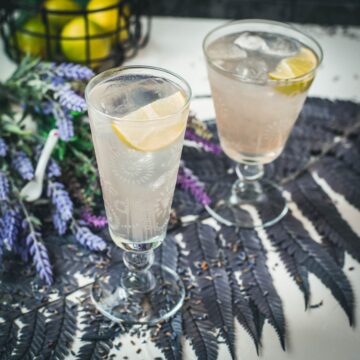 Two beautiful glasses with flower engraving filled with lavender Moscow Mule, arranged on dark blue purple leaves.