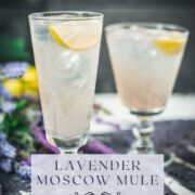 Two beautiful glasses with flower engraving filled with lavender Moscow Mule, arranged on dark blue purple leaves.