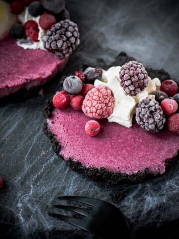 Hauntingly delicious mini Oreo tarts filled with a berry mix ganache, garnished with a creepy gummy eye, frozen berries and whipped cream. Placed on a black plate covered with spider webs.