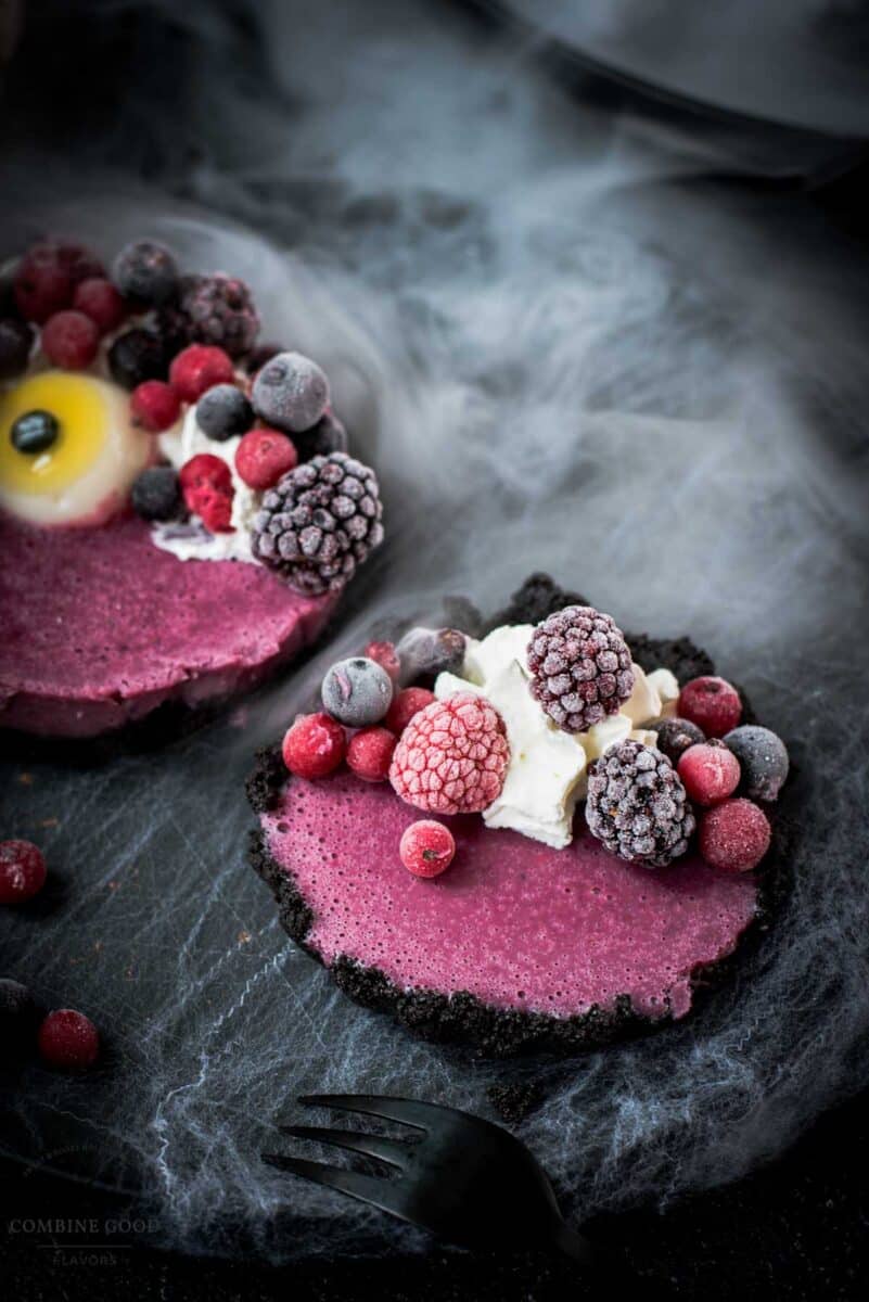 Hauntingly delicious mini Oreo tarts filled with a berry mix ganache, garnished with a creepy gummy eye, frozen berries and whipped cream. Placed on a black plate covered with spider webs.