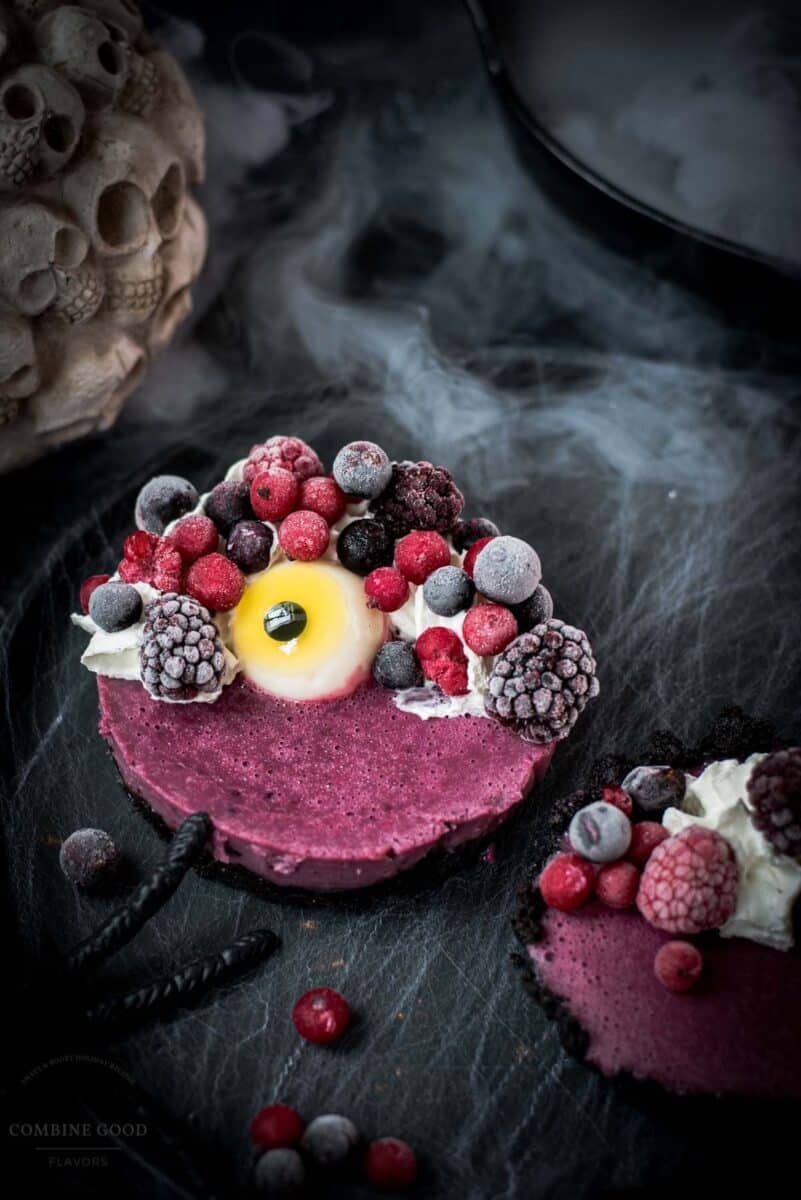 Hauntingly delicious mini Oreo tarts filled with a berry mix ganache, garnished with a creepy gummy eye, frozen berries and whipped cream. Placed on a black plate covered with spider webs.