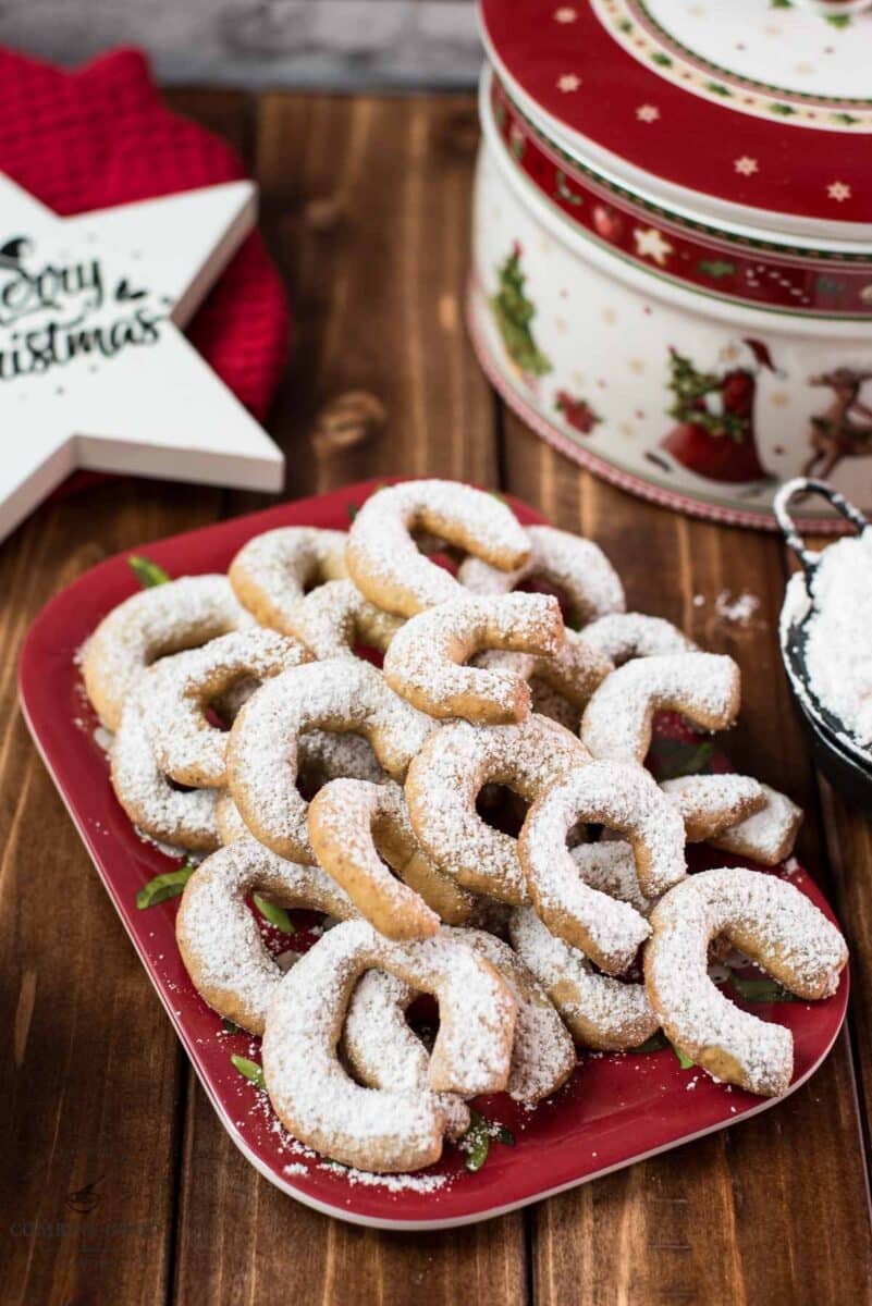 Delicious traditional Austrian vanilla crescents stacked on christmassy plate, placed next to a Christmass cookie jar.