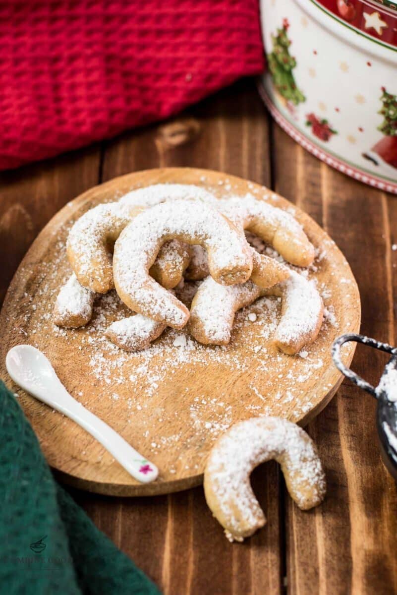 Delicious traditional Austrian vanilla crescents stacked on wooden plate, placed next to a Christmass cookie jar.