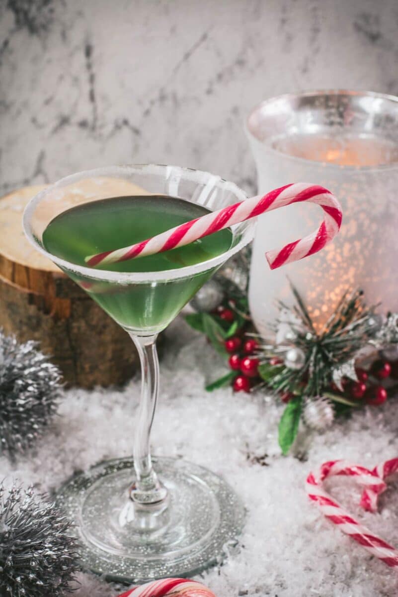 Festive cranberry peppermint martini in sugar rimmed martini glass. Garnished with a candy cane, placed on snowy ground.