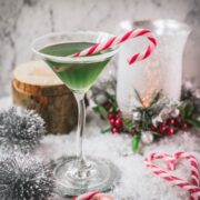 Festive cranberry peppermint martini in sugar rimmed martini glass. Garnished with a candy cane, placed on snowy ground.