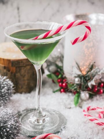 Festive cranberry peppermint martini in sugar rimmed martini glass. Garnished with a candy cane, placed on snowy ground.