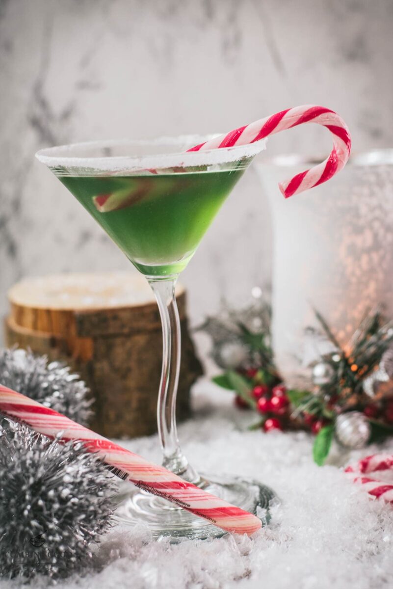 Festive cranberry peppermint martini in sugar rimmed martini glass. Garnished with a candy cane, placed on snowy ground.