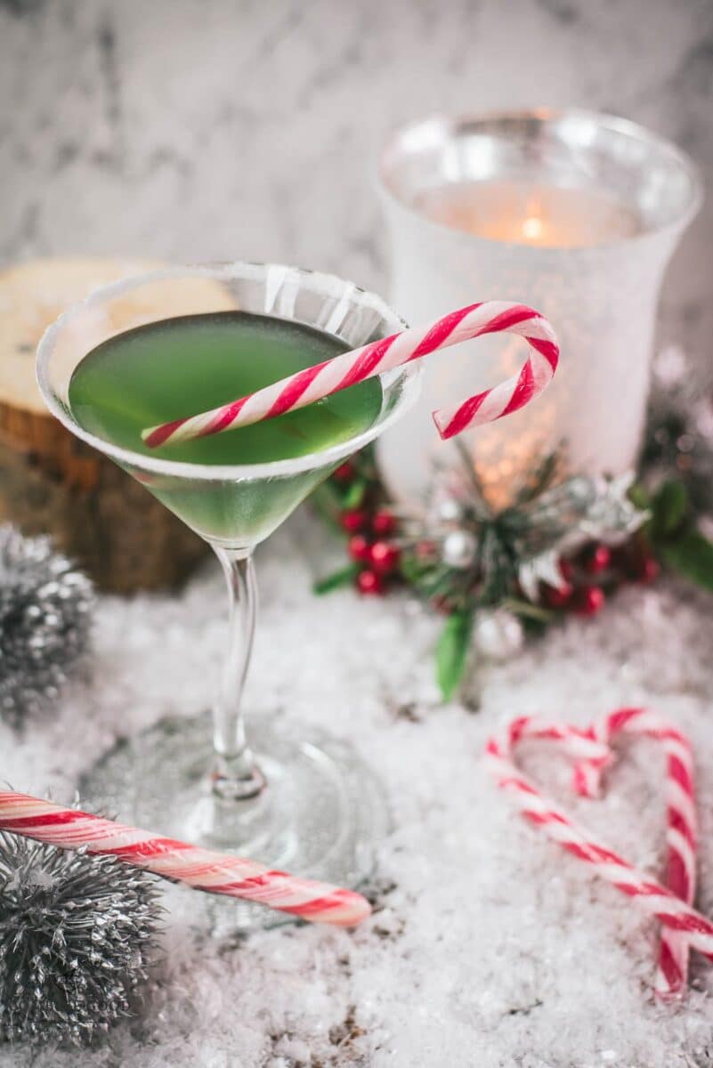 Festive cranberry peppermint martini in sugar rimmed martini glass. Garnished with a candy cane, placed on snowy ground.