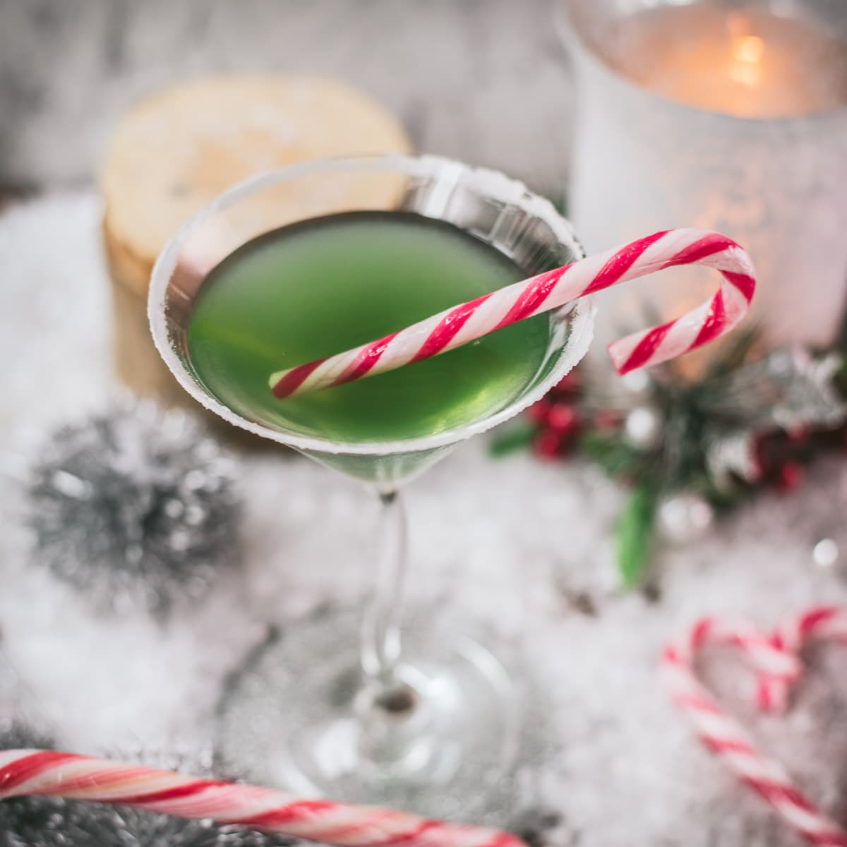 Festive cranberry peppermint martini in sugar rimmed martini glass. Garnished with a candy cane, placed on snowy ground.