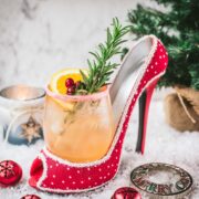 Festive vodka cranberry spritzer, garnished with rosemary twig, orange slice, and cranberries, served in a glass that is placed in a christmassy red high heel.