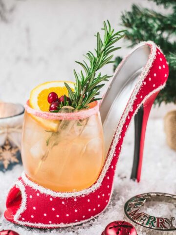 Festive vodka cranberry spritzer, garnished with rosemary twig, orange slice, and cranberries, served in a glass that is placed in a christmassy red high heel.