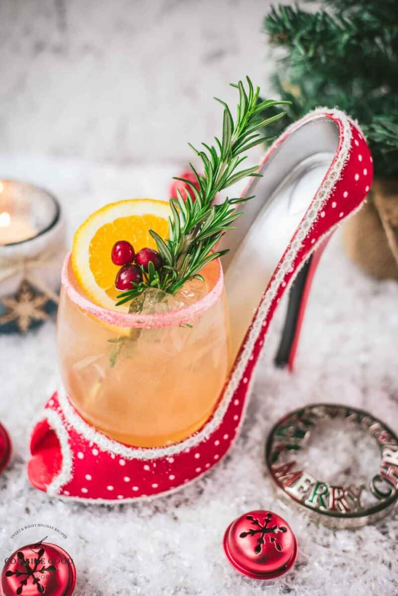 Festive vodka cranberry spritzer, garnished with rosemary twig, orange slice, and cranberries, served in a glass that is placed in a christmassy red high heel.