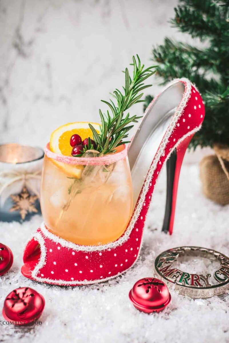 Festive vodka cranberry spritzer, garnished with rosemary twig, orange slice, and cranberries, served in a glass that is placed in a christmassy red high heel.