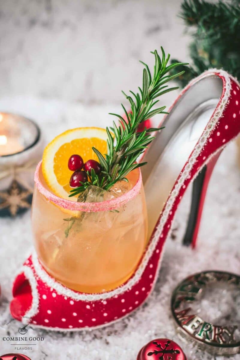 Festive vodka cranberry spritzer, garnished with rosemary twig, orange slice, and cranberries, served in a glass that is placed in a christmassy red high heel.