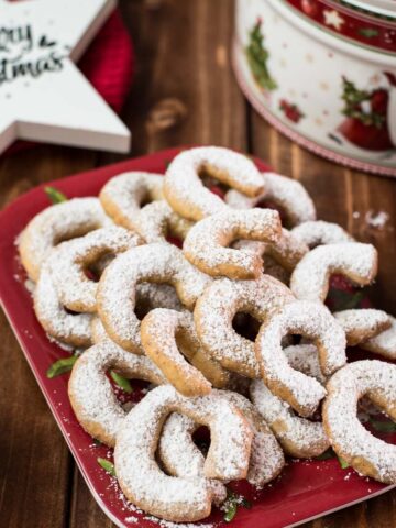 Delicious traditional Austrian vanilla crescents stacked on christmassy plate, placed next to a Christmass cookie jar.