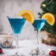 Gorgeous cocktail glasses filled with a stunning blue Christmas cocktail, garnished with an orange slice. Placed on mirrored coasters on snowy ground.