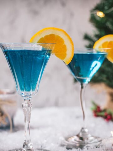 Gorgeous cocktail glasses filled with a stunning blue Christmas cocktail, garnished with an orange slice. Placed on mirrored coasters on snowy ground.