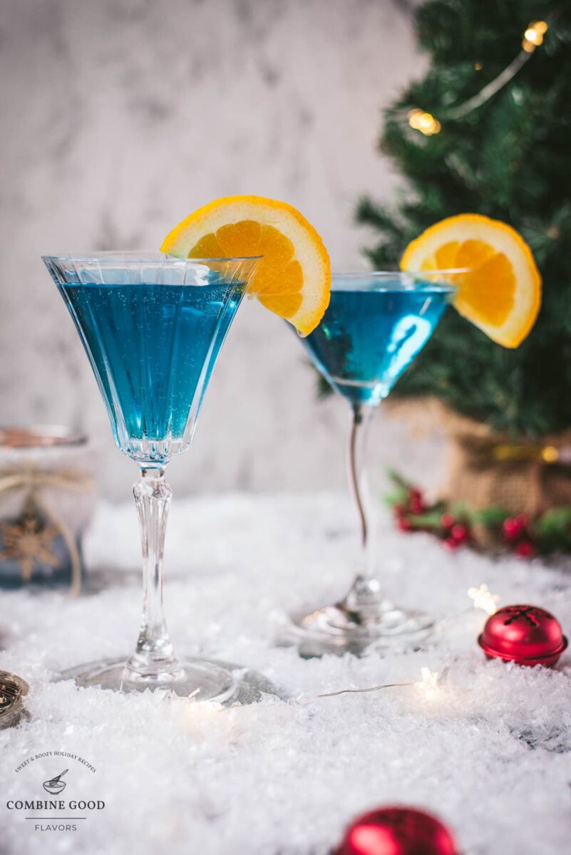 Gorgous cocktail glasses filled with a stunning blue Christmas cocktail, garnished with an orange slice. Placed on mirrored coasters on snowy ground.