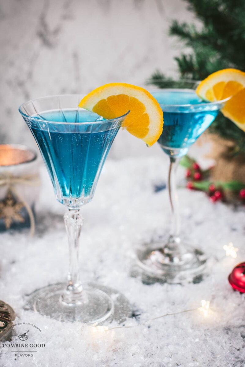 Gorgous cocktail glasses filled with a stunning blue Christmas cocktail, garnished with an orange slice. Placed on mirrored coasters on snowy ground.