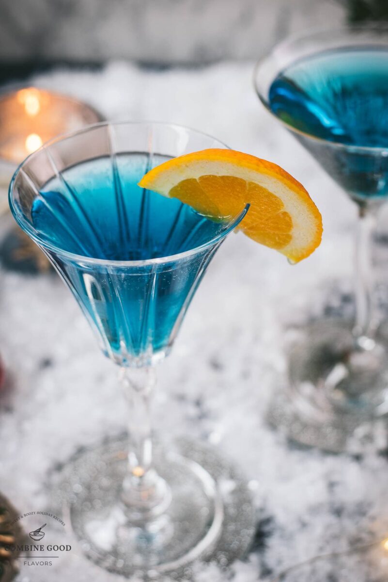 Gorgous cocktail glasses filled with a stunning blue Christmas cocktail, garnished with an orange slice. Placed on mirrored coasters on snowy ground.