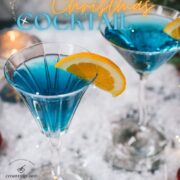 Gorgeous cocktail glasses filled with a stunning blue Christmas cocktail, garnished with an orange slice. Placed on mirrored coasters on snowy ground.