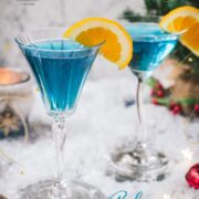 Gorgeous cocktail glasses filled with a stunning blue Christmas cocktail, garnished with an orange slice. Placed on mirrored coasters on snowy ground.